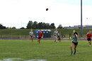 High school girls playing flag football on a grass field at a school. Several players are wearing orange and black jerseys. The others are wearing gray and green jerseys. 