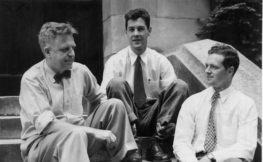 A historic black-and-white photograph of sexologist Alfred Kinsey wearing a bowtie, sitting on stone steps outside with two male members of his research team.