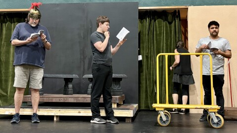 People on a stage with roman helmet and pillar seats behind them.