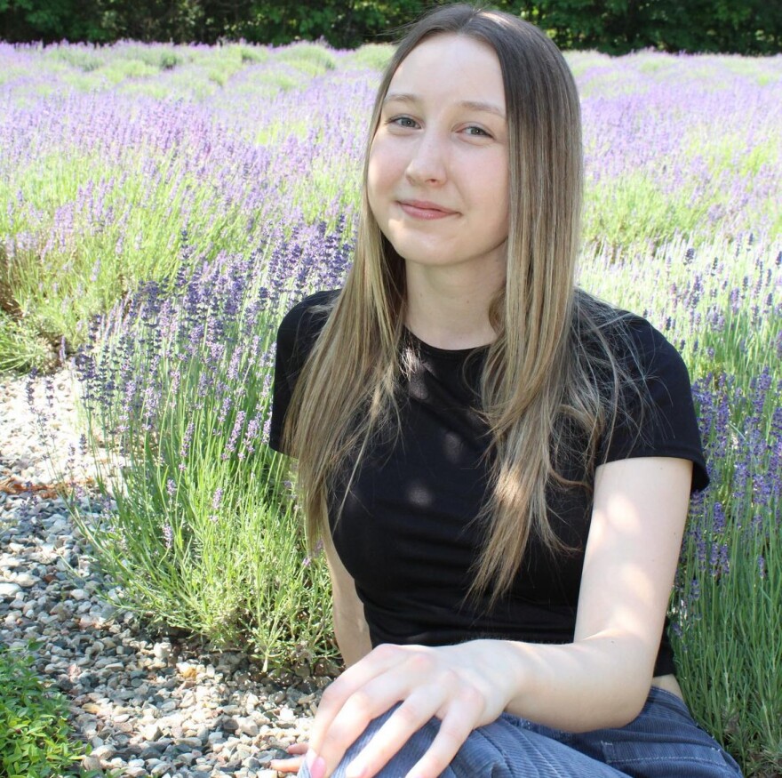 Rachel Noreen young white woman with blonde hair poses in a field of lavender. She's wearing jeans and a black t-shirt. 