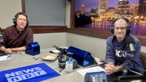 Two smiling men wearing headphones sit at a table in a radio talk studio: the man at left has short brown hair and is wearing a maroon button-down shirt; the man at right has short dark hair and is wearing glasses and a navy blue hoodie with "WXXI" in white lettering