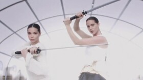 Two women dressed in white wield samurai swords while looking directly at the camera and posing against an illuminated white background.