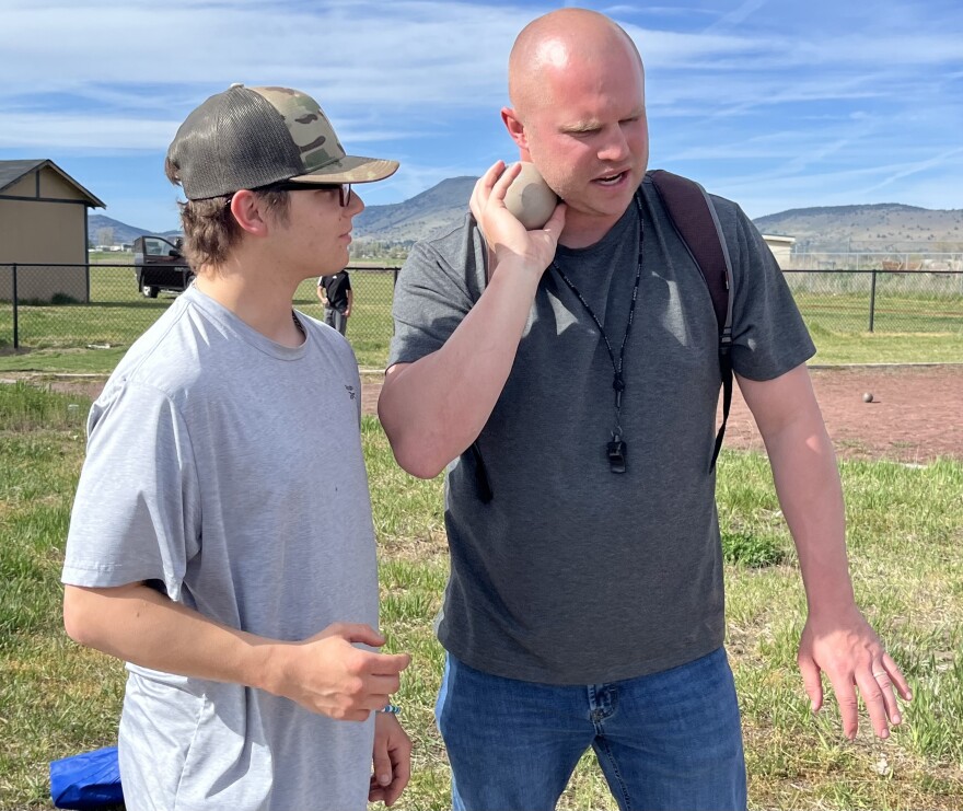 A middle aged, bald man holds a shot put next to a teenager in a gray t shirt with glasses. Behind them is a green field and blue sky. 