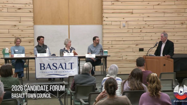 Four people sit at a table, facing a small seated audience while listening to a man speak at a podium