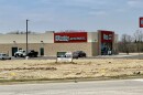 A real estate sign stands on an empty lot in front of a brown building occupied by an auto parts business, with cars parked alongside. 