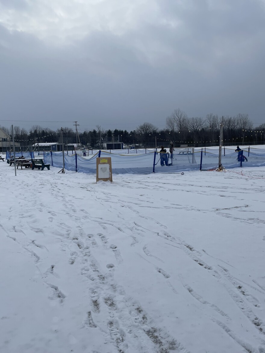 A photo of footsteps in snow leading top an ice rink surrounded by string lights and blue netting on a grey, winter day.