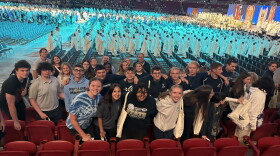 Several dozen high school students pose for a photo in an indoor arena as a gathering of men in white robes walk in a procession at a Catholic church gathering.