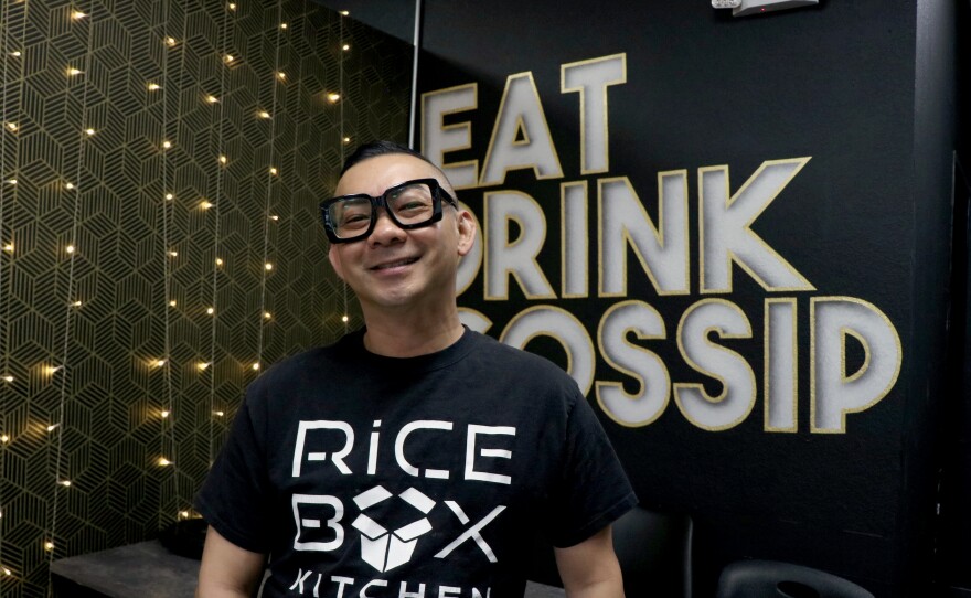 Perapol Damnernpholkul smiles while standing in front of a painted sign behind him that says “Eat, Drink, Gossip.”