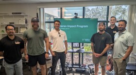 Group of men stand in room in front of screen showing presentation of veteran impact program 