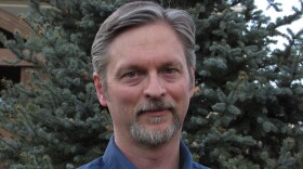 Collin Irish, a Colorado writer, wears a blue button down shirt and stands in front of an evergreen tree