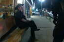 A woman in a cowboy hat sits on a bench and talks to a cop. Cop cars are in the background.