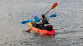 two people sit in a two-person orange kayak paddling out to sea. They have a box attached to the back of the kayak