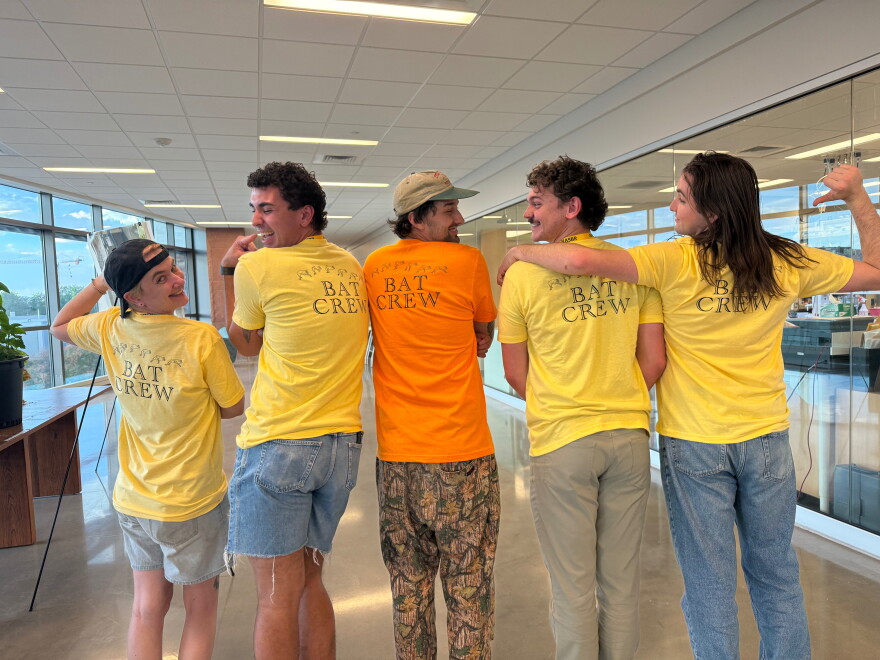Several students stand in a line with backs toward the viewer. They're all wearing yellow and orange shirts that say "BAT CREW" with a bat logo on it. Some point back at the shirt, and some are looking back and smiling.