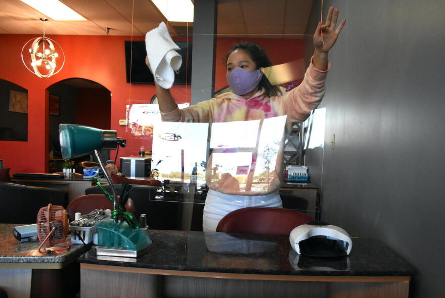A younger worker at a nail salon cleans a plexiglass shield being used to prevent the spread of COVID-19.