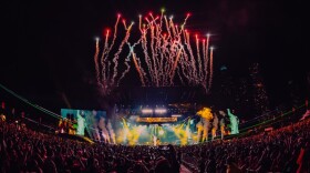 Fireworks go off above a stage where an artist performs in front of a large audience.