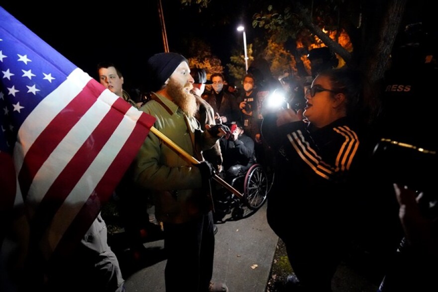 Two people face each other in a nighttime protest scene. One is holding an American flag.