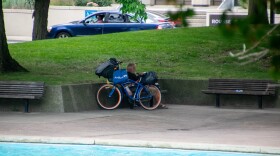 A man sits behind a bike with multiple bags strapped to it, avoiding the sun in Freimann Square.