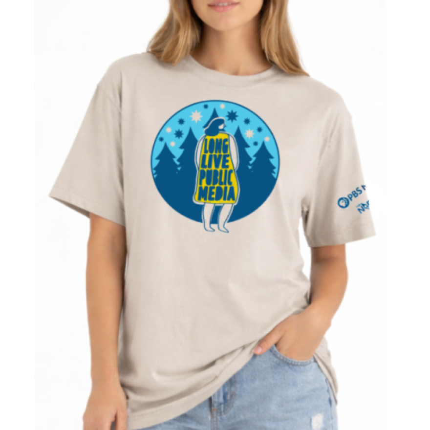 A beige t-shirt with a circular design featuring a drawing of a woman facing away wearing a yellow dress that says Long Live Public Media. In the background are dark pine trees and stars against a light blue sky.