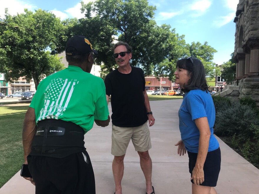 Willie Hudspeth, in a green T-shirt, has his back to the camera as he shakes hands with Gene Ross, a man wearing sunglasses. Barbara Ross talks off to the side.