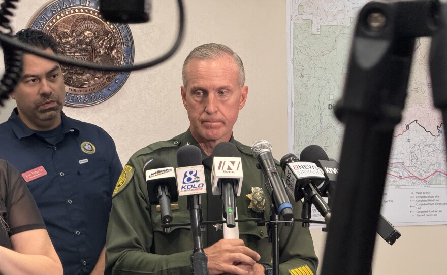 A sheriff standing up with several microphones pointed toward him. He is looking away from the camera.