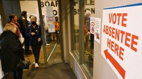 People stand in line to the left while a woman exits a door. To the right is a sign that reads "Vote Absentee Here."