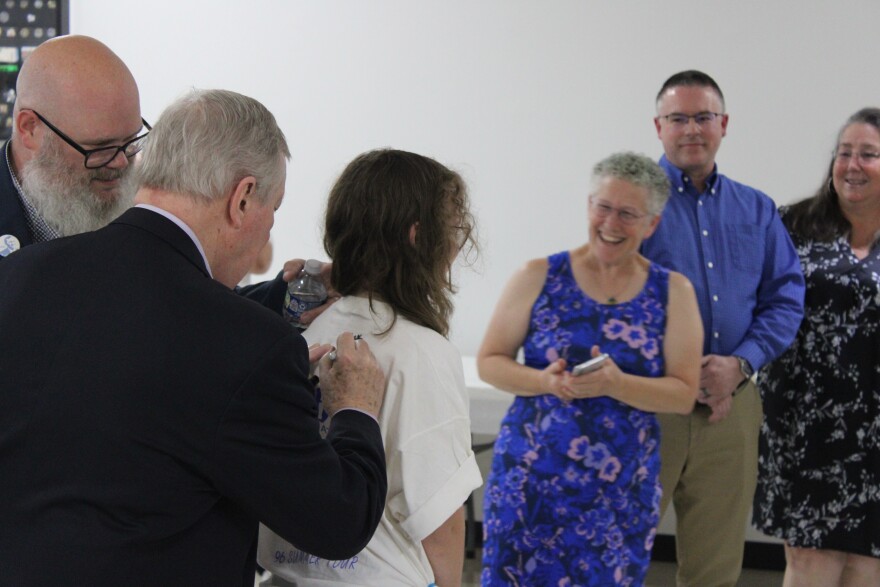 Dick Durbin signs a t-shirt on a child's back, several observers smile behind them. 