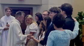 A man wearing white religious robes holds hands with a young woman surrounded by other people in a church.