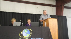 A business executive speaks at a podium next to a table set up for a public meeting. 