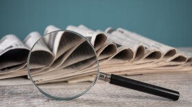A magnifying glass and newspapers stacked on a wooden table on a blue background