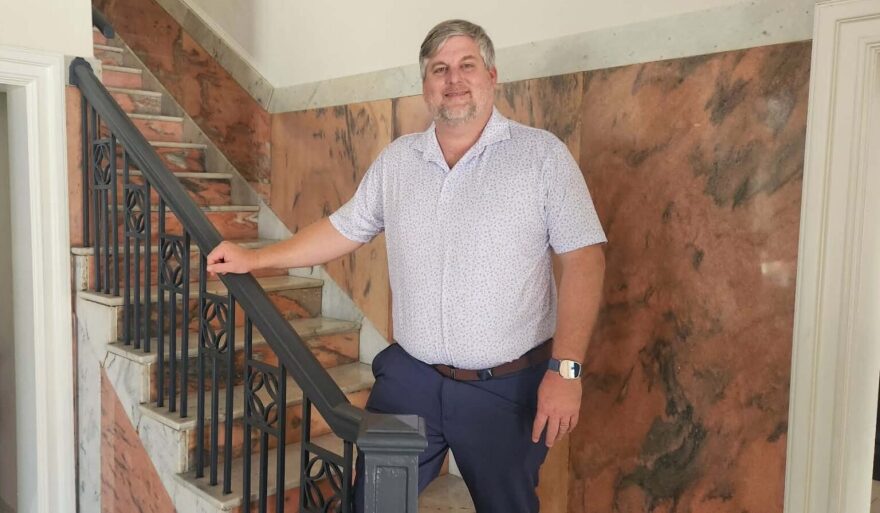 Man with grey hair wears polo shirt and stands next to staircase 
