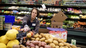 A woman wearing an apron and name tag at a grocery store is sorting through produce which includes potatoes, squash and apples.