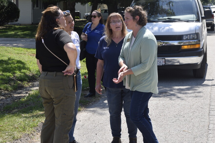 Four women standing next to each other in a semi circle on a residential street and in front of a white van.