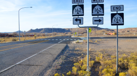 Road signs off a rural street in an arid area. There are a few buildings in the distance and mountains beyond that.