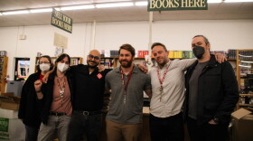 Six people stand in a line in front of a counter and bookshelves. A sign above them reads "Sell your books here"