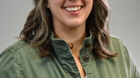 Woman with medium-length hair wearing a green jacket over a white shirt, smiling at the camera on a gray background.