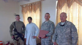 An image shows three men dressed in camouflaged standing with another man dressed in all white inside a room with gold curtains on the windows behind them.