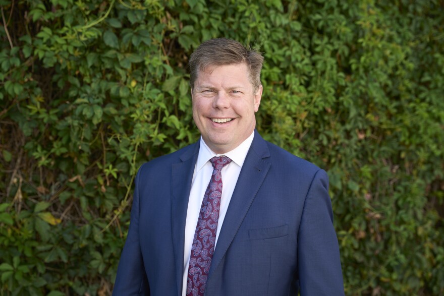 A white man in a blue suit smiles at the camera with a green ivy wall behind them.