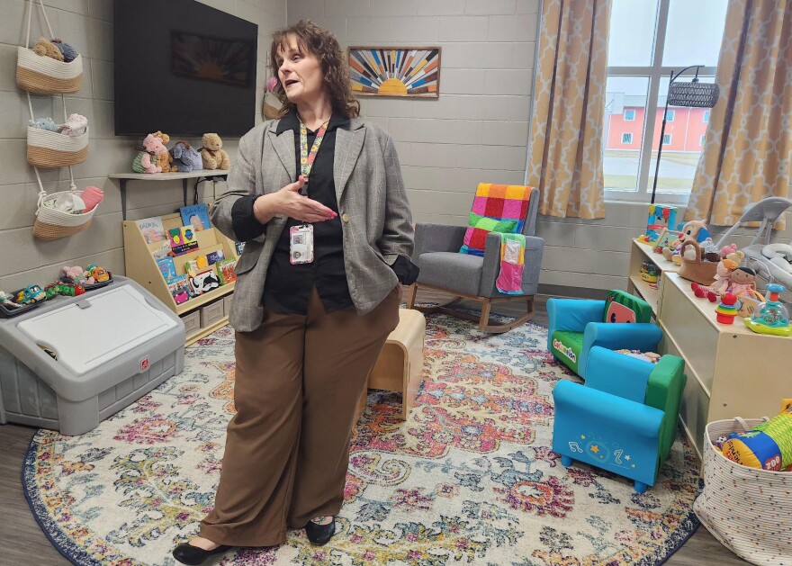 Kim Perkins wears a gray blazer and brown slacks. She stands in the middle of a daycare center - with a colorful rug on the floor and toys all around.