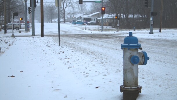 A blue and silver fire hydrant is in the right side of the frame, snow surrounds it. In the background are traffic lights and an intersection. The roads are snow covered.