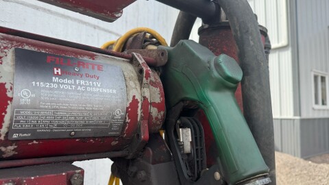A green gas handle attached to a large, white, cylindrical gas tank is exposed to the open air. 