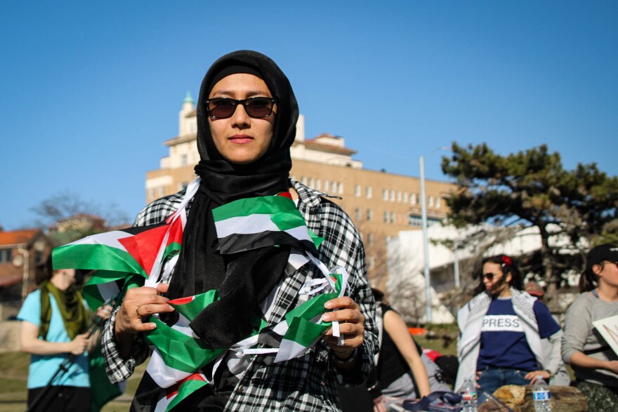 a woman with a string of Palestinian flags around her neck stands and looks solemnly at the camera