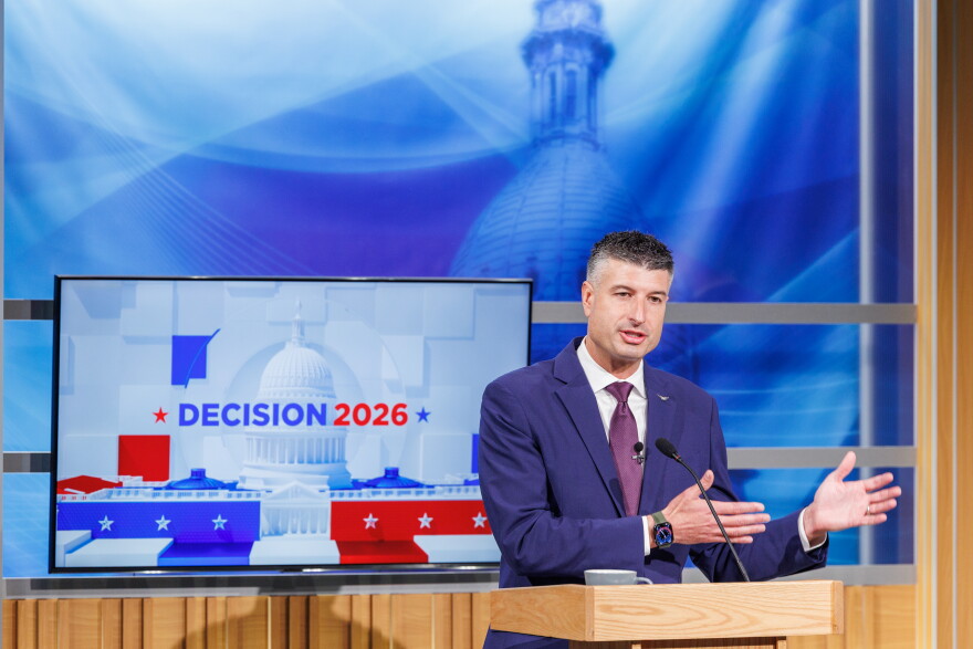 A man in a suit speaks at a podium in front of a TV that reads "Decision 2026"