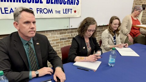 A man and two women sit at a table with a blue tablecloth. The man on the left is holding a microphone.