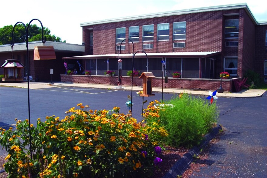 A spring flower garden with birdfeeders hanging from poles, set in a parking lot, is in the foreground of an image of a red brick, two-story building.