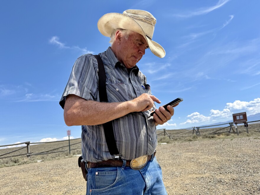 A rancher in a cowboy hat an suspenders looks at his phone on a sunny day.