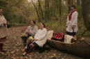 Four individuals smile and laugh while in a forest near a body of water. Two of the individuals are leaning against a boat, while the other two are standing. 