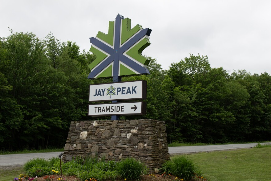 A sign in the shape of a maple leaf painted green and blue and the words "Jay Peak Tramside" sits by a road in summer.