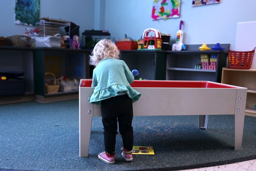 A toddler, wearing a light green blouse, black pants and pink sneakers, plays in a toy room.