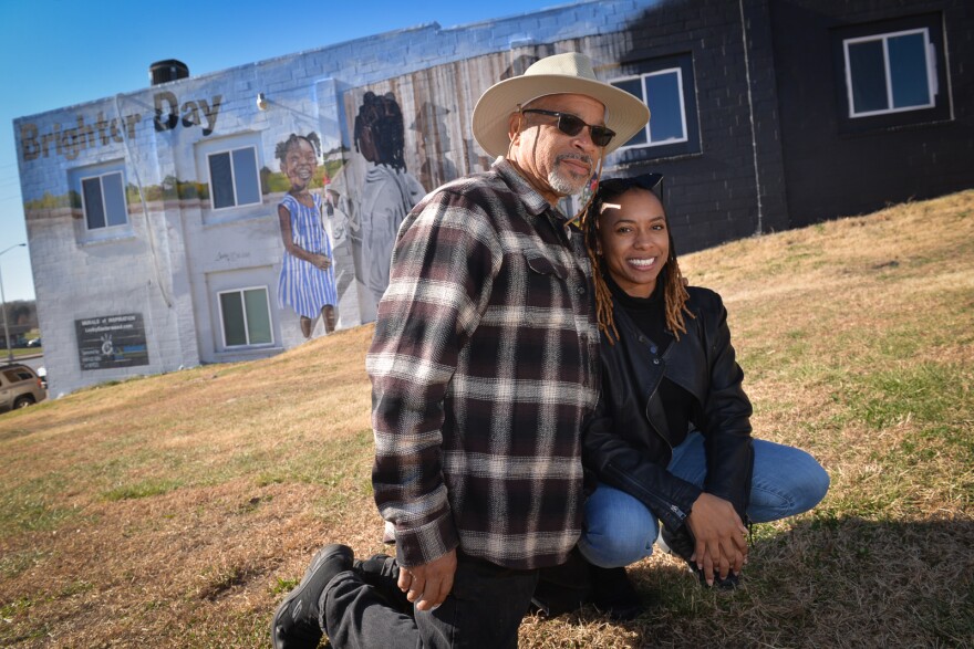 Man wearing a hat and plaid shirt kneeling on ground with woman in black leather jacket squatting beside him. There is a building in background with a mural titled "Brighter Day" behind them. They are father and daughter.