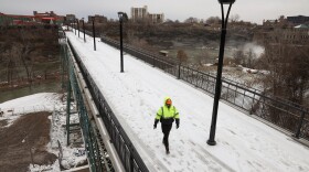 A construction worker walks along the snow-covered deck of the Pont de Rennes pedestrian bridge over the Genesee River gorge. Genesee Brewery is seen in the background, as well as the spray of the falls.